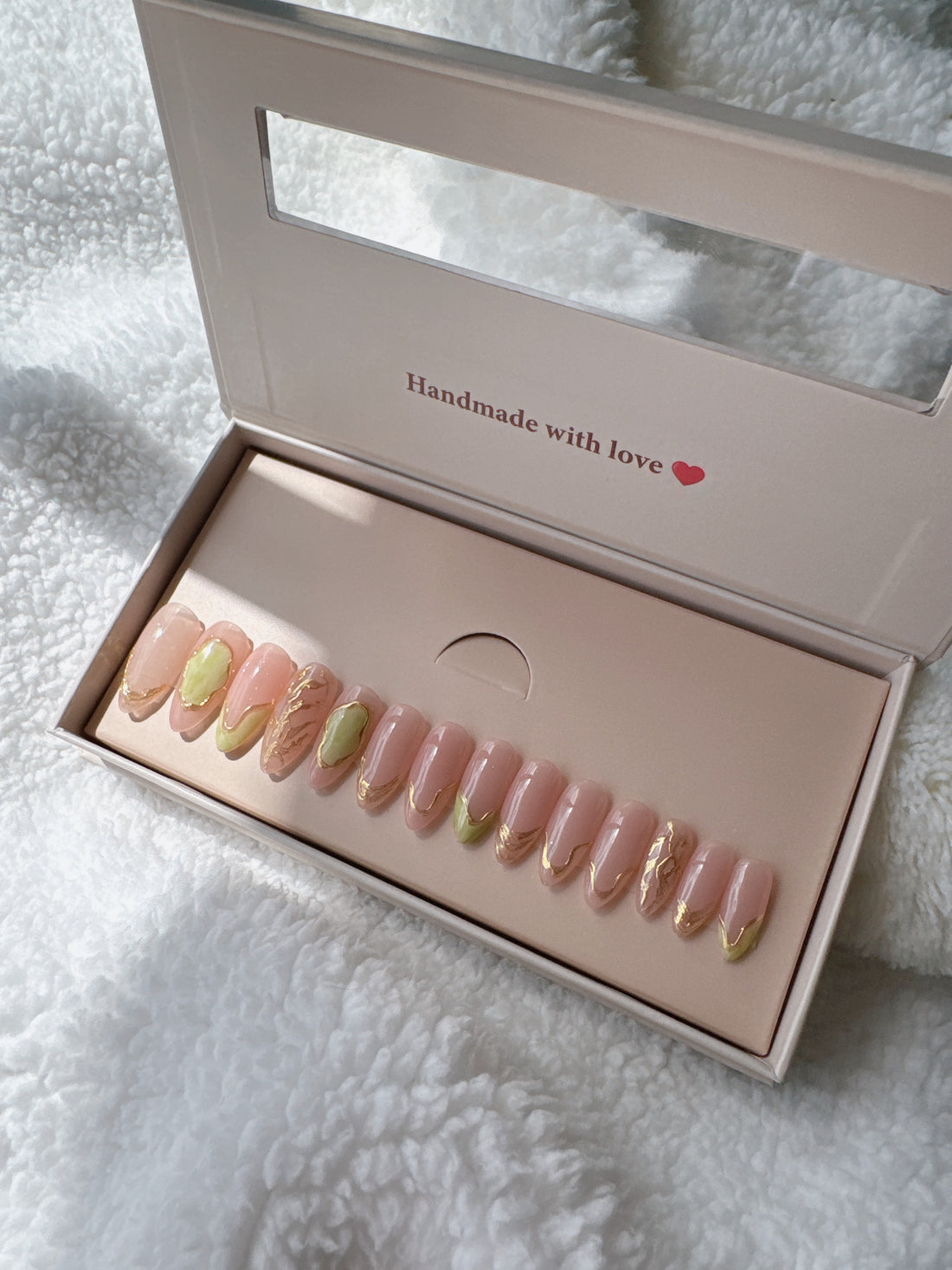 Box of nude and gold press-on nails on soft white textured fabric