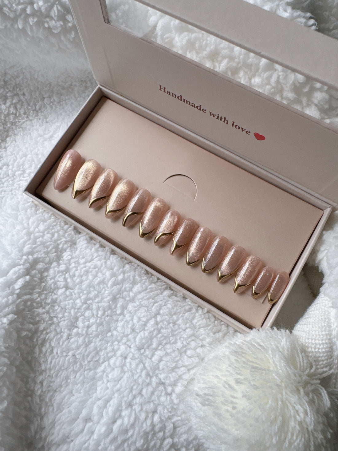 Box of nude and gold press-on nails on a soft white textured fabric background