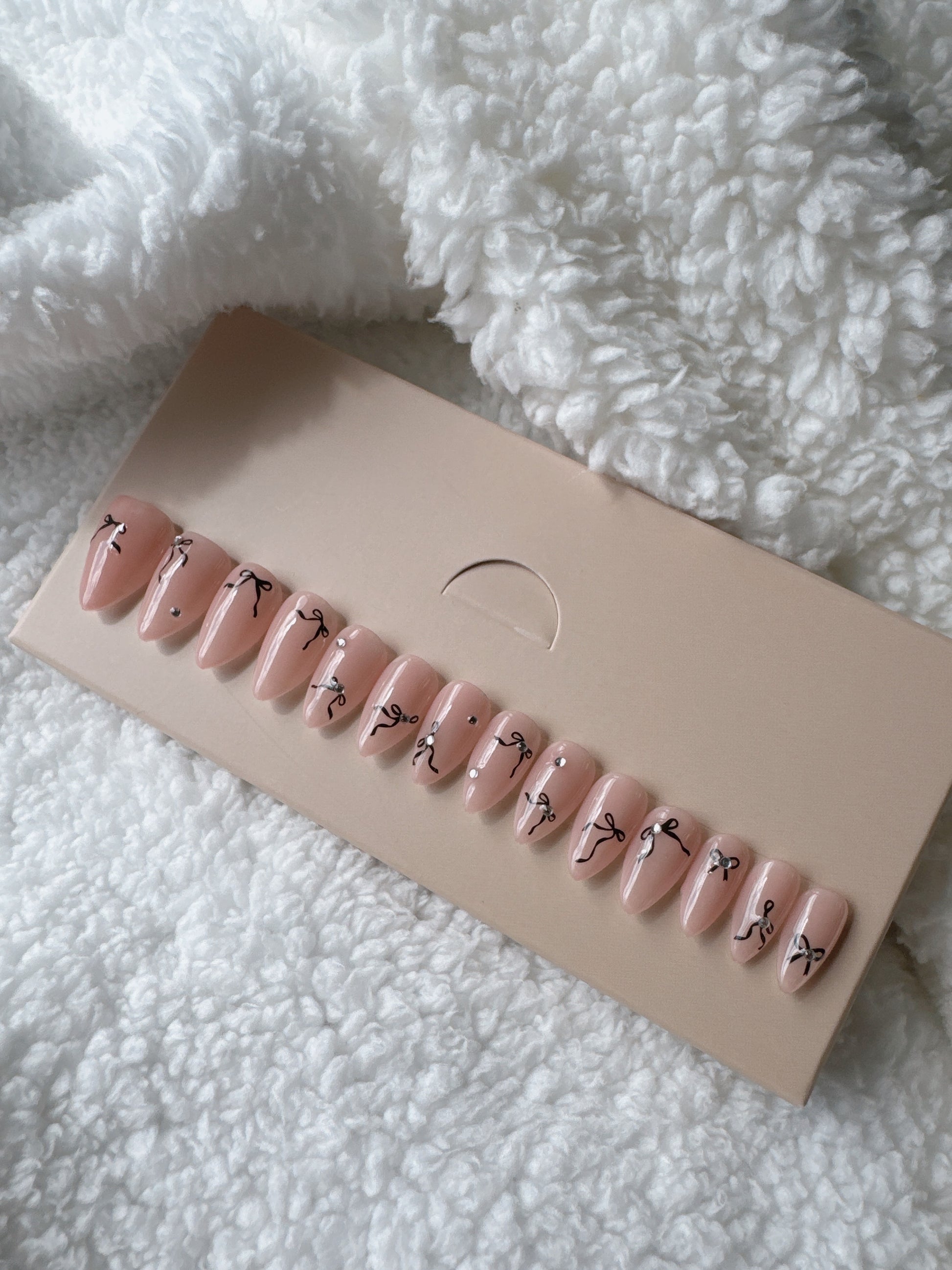 Almond-shaped nude press-on nails with black bow designs and rhinestone accents on display card