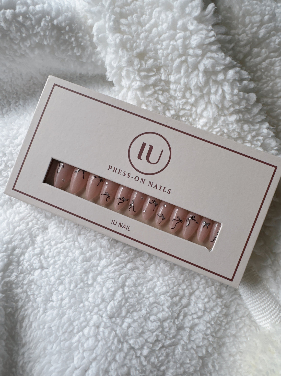 Box of nude press-on nails with black bow designs on white textured fabric