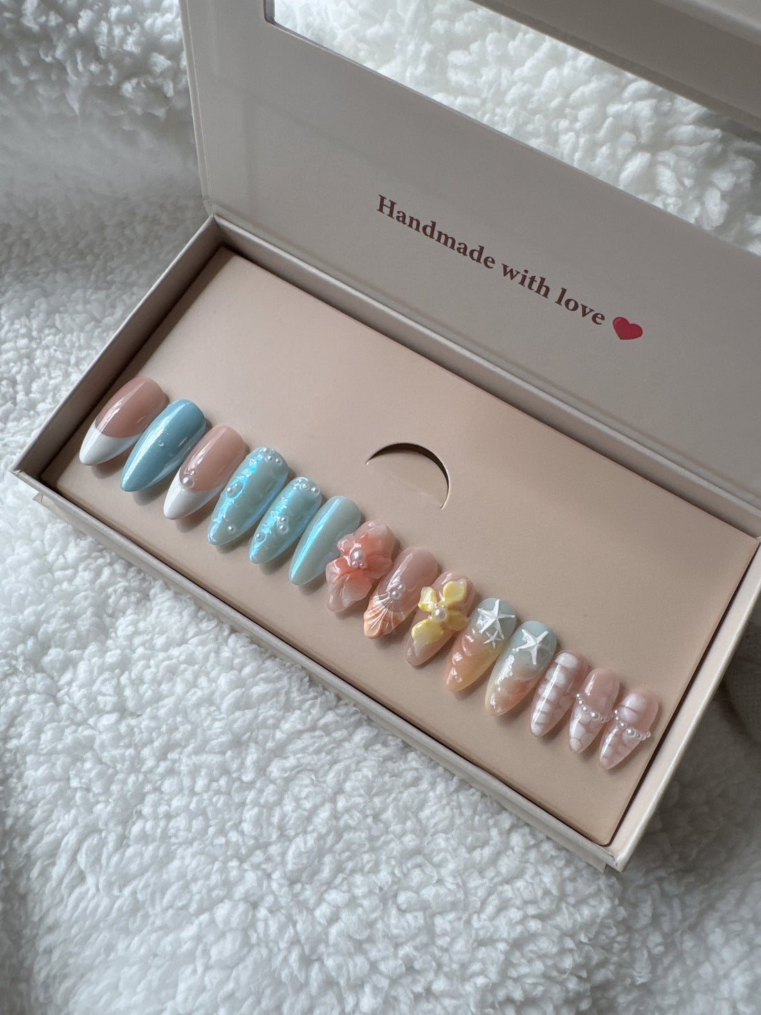 Box of colorful press-on nails with nail art designs on a white fluffy fabric background