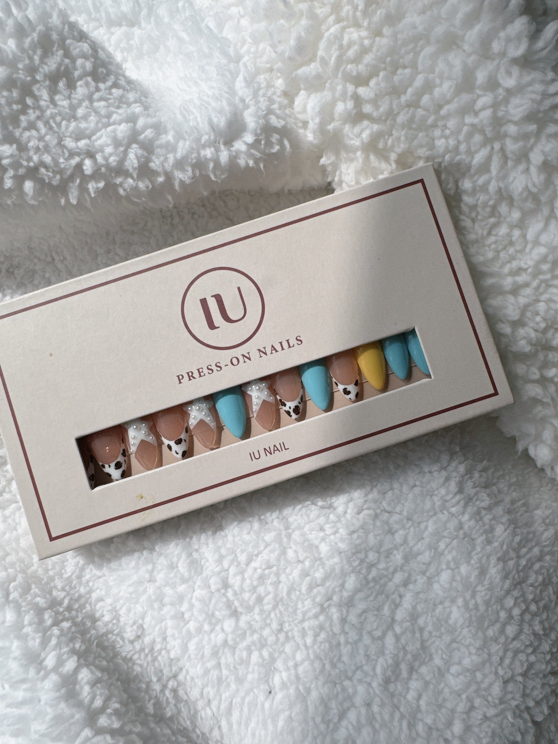 Box of multicolor press-on nails with cow print and pearl accents on white fabric