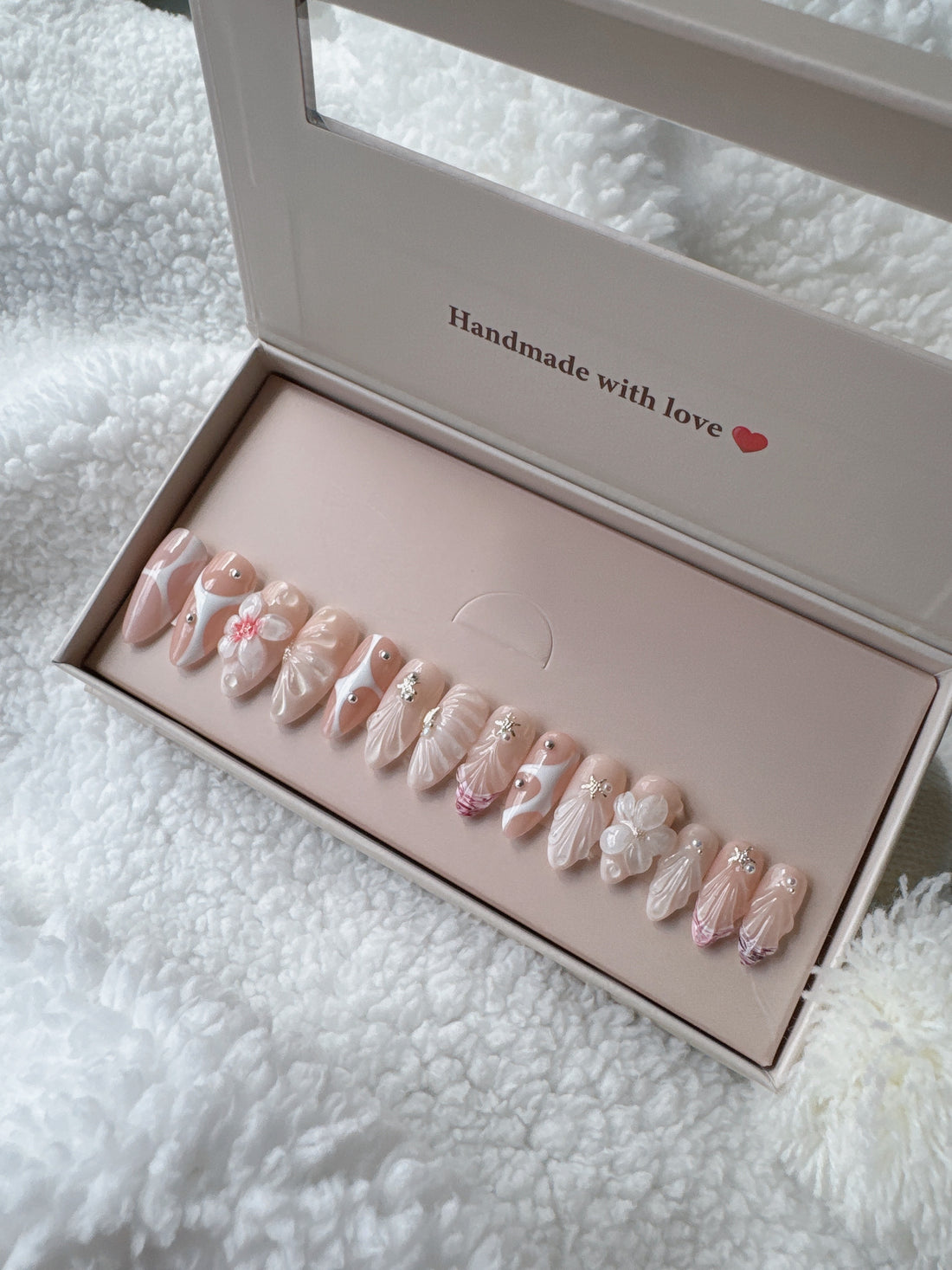 Box of pink and white press-on nails with floral and jeweled designs on white fabric