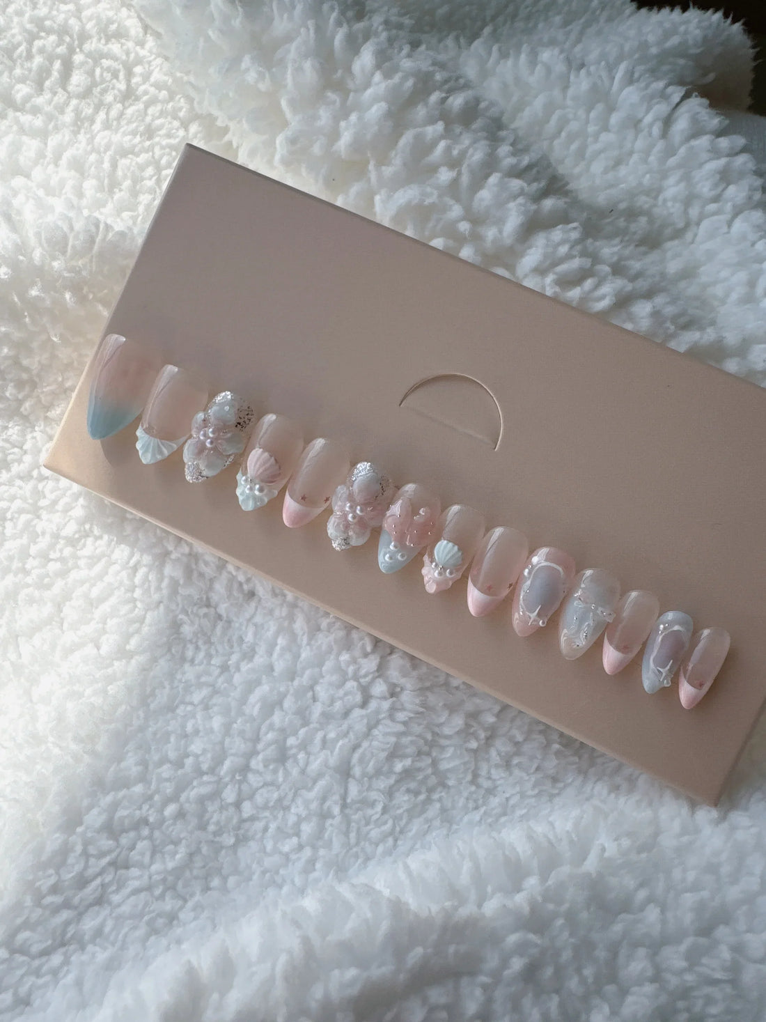 Box of pastel pink and blue press-on nails with 3D floral designs on soft white fabric.