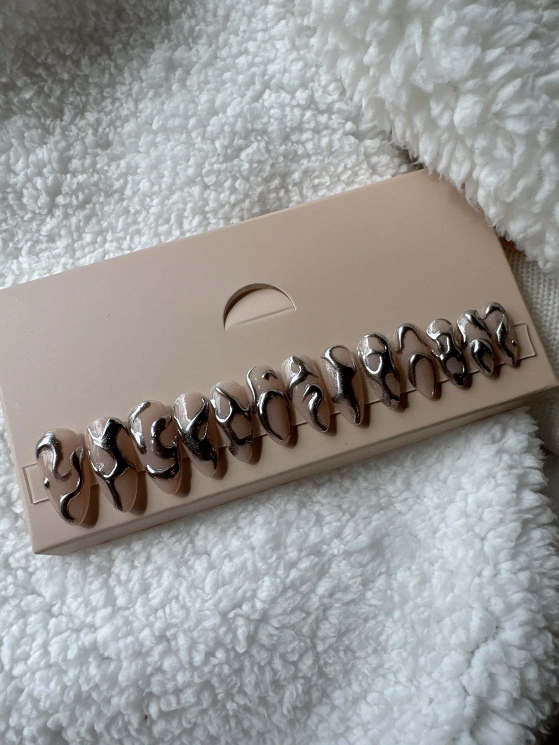 Glossy nude press on nails with abstract chrome silver designs on a display card, on soft fabric background.