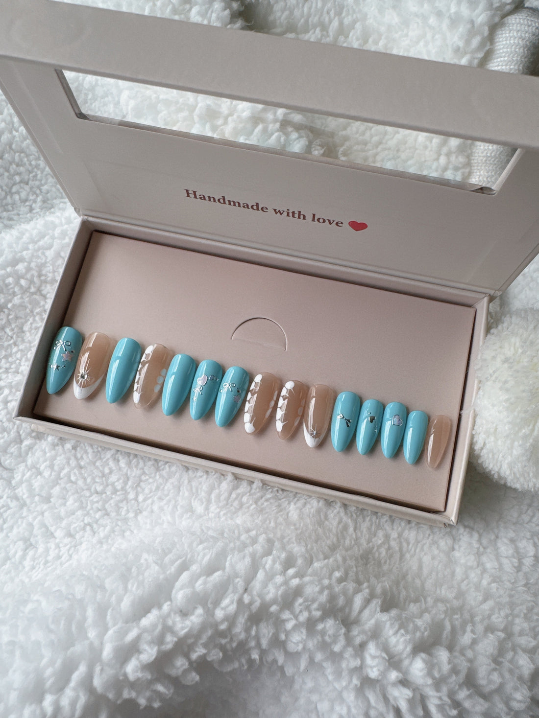 Blue and nude press-on nails with white floral designs in a box on white textured fabric.