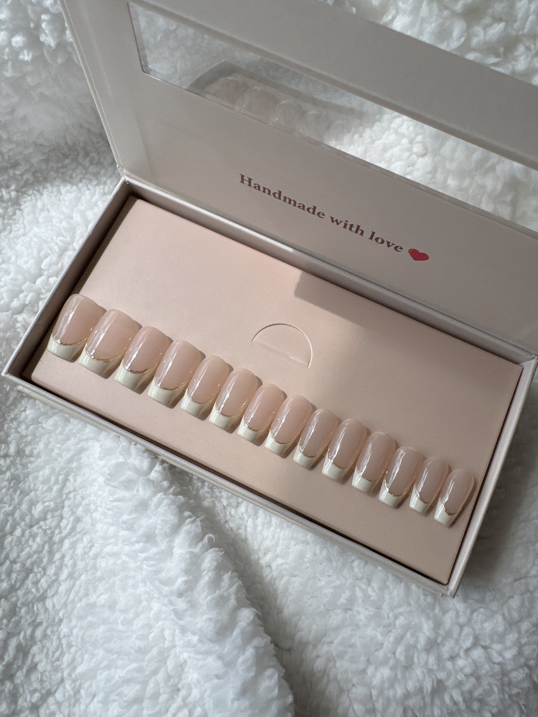 Box of nude French tip press-on nails on a soft white textured fabric background