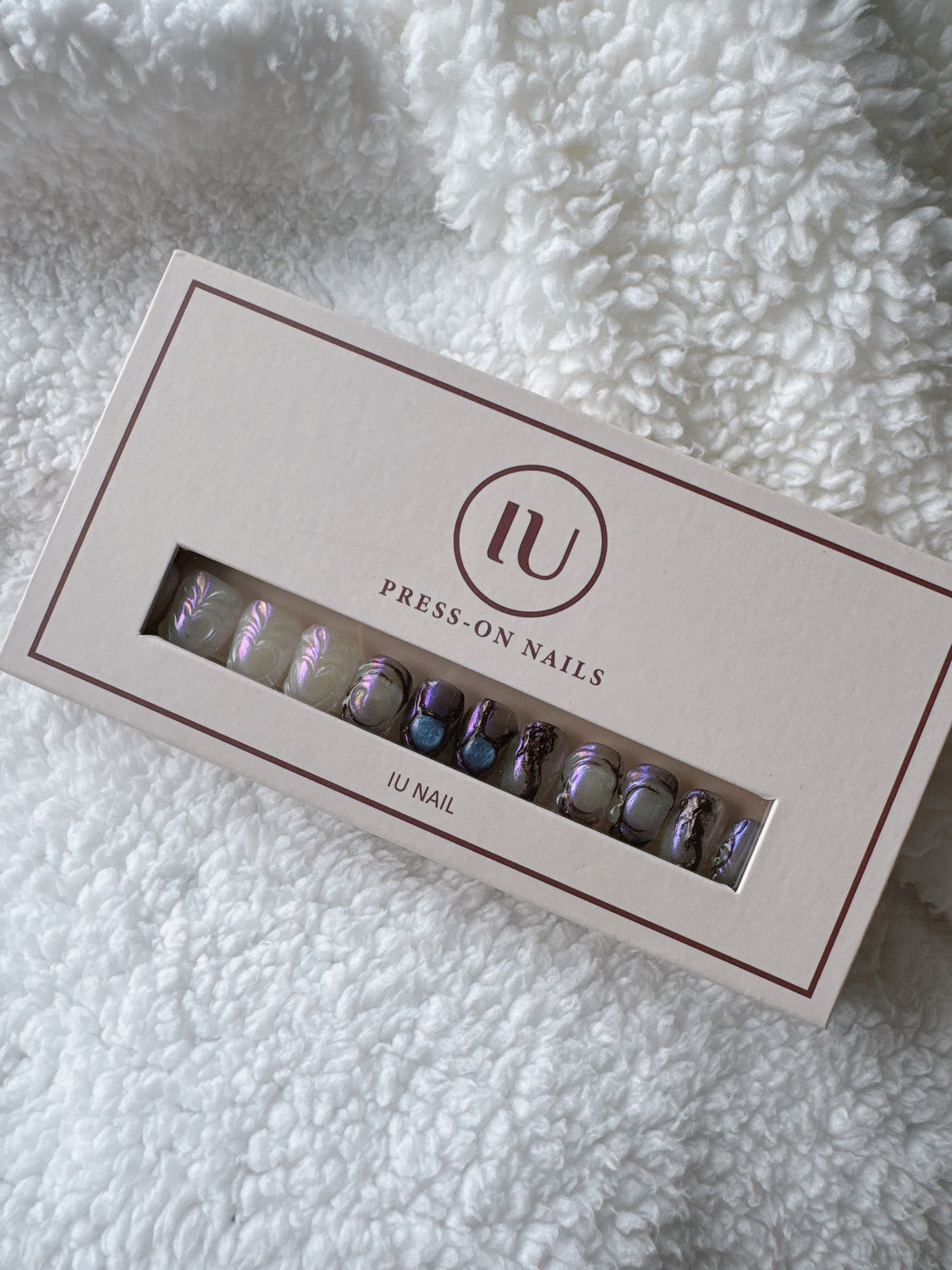 Iridescent press-on nails set in a beige box on a soft white textured blanket