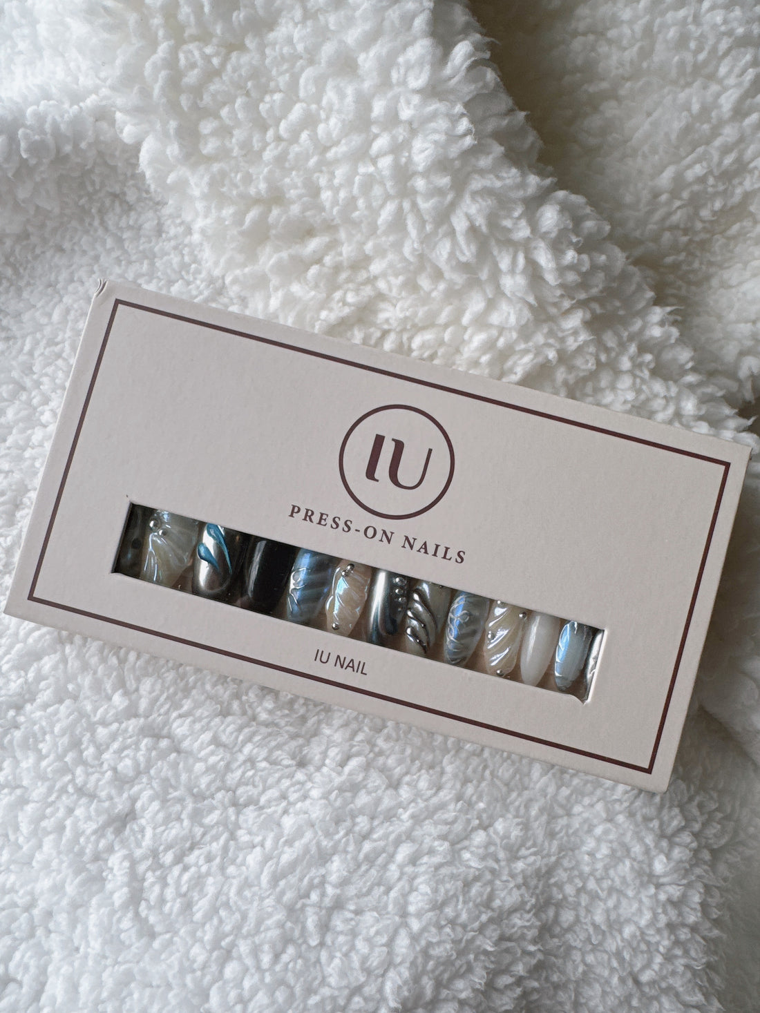 Box of IU press-on nails with marble and metallic designs on a white fluffy fabric