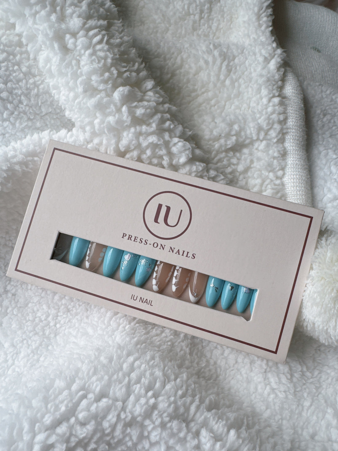 Blue and nude press-on nails with white floral designs in a box on white textured fabric.