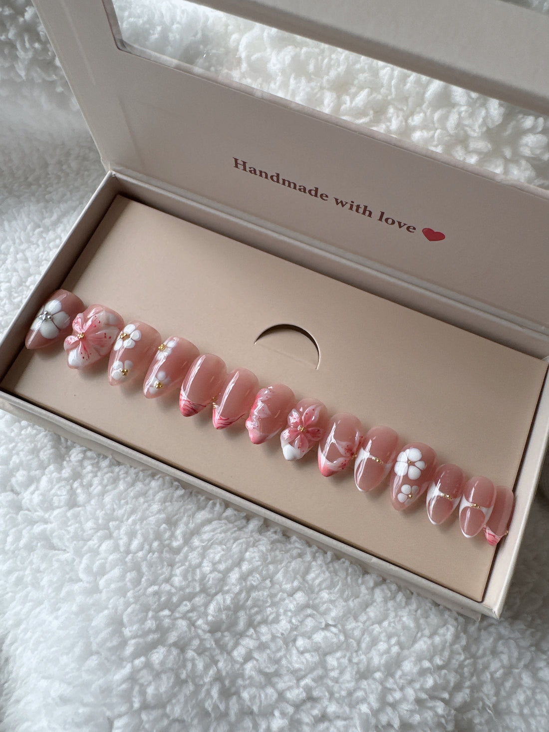 Box of pink floral press-on nails with gold accents on white textured fabric background