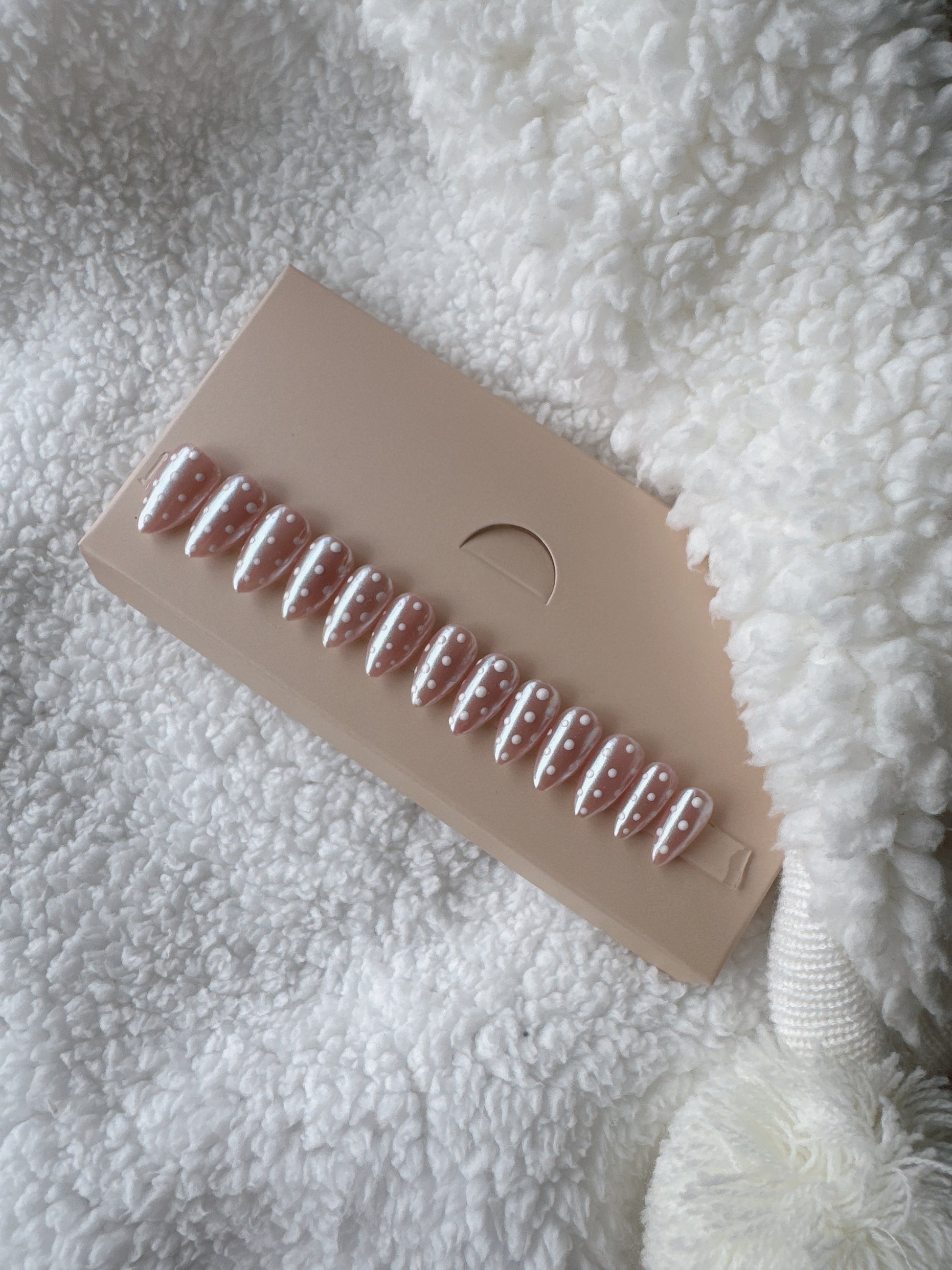 Almond-shaped nude press on nails with white polka dot design on beige card, on soft white fabric.