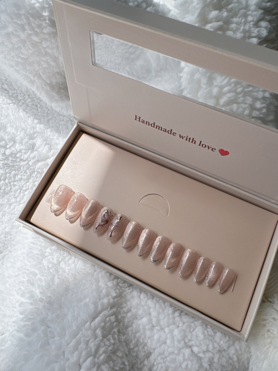 Box of nude press-on nails with silver tips and floral accents on white textured fabric