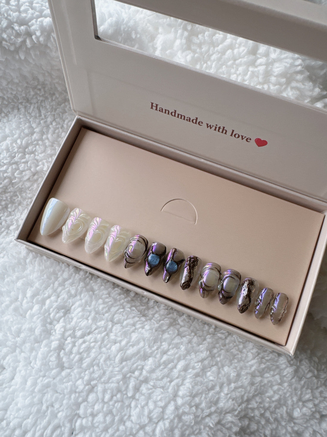 Iridescent press-on nails set in a beige box on a soft white textured blanket