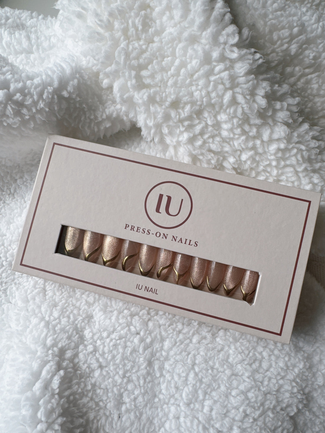 Box of nude and gold press-on nails on a soft white textured fabric background