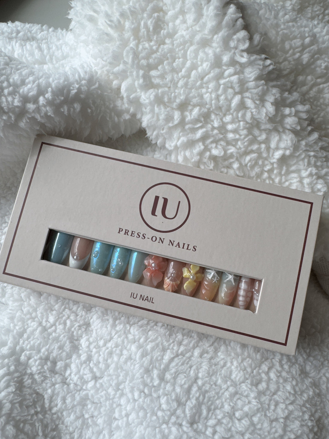 Box of colorful press-on nails with nail art designs on a white fluffy fabric background