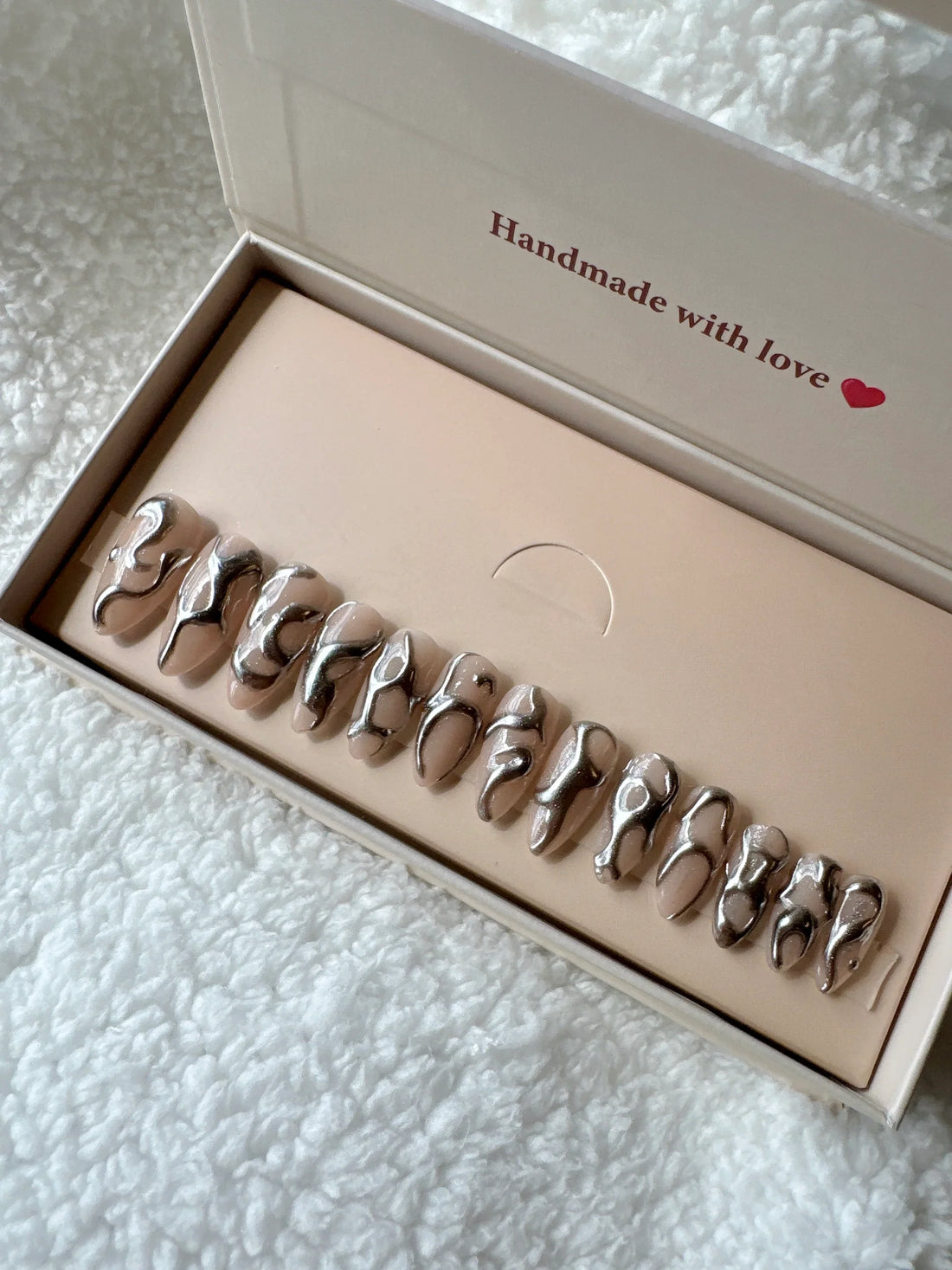 Glossy nude press on nails with abstract chrome silver designs on a display card, on soft fabric background.