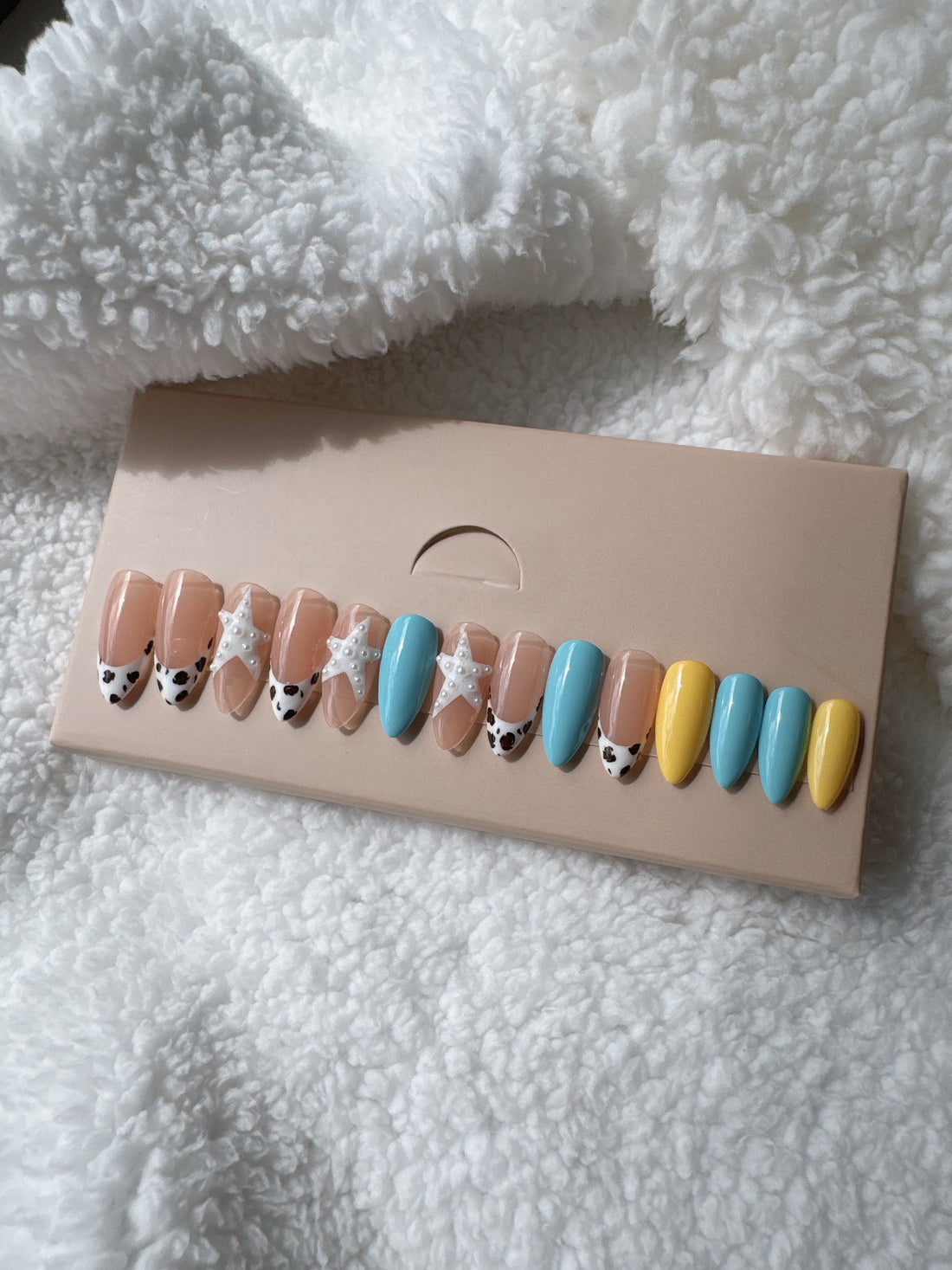 Box of multicolor press-on nails with cow print and pearl accents on white fabric