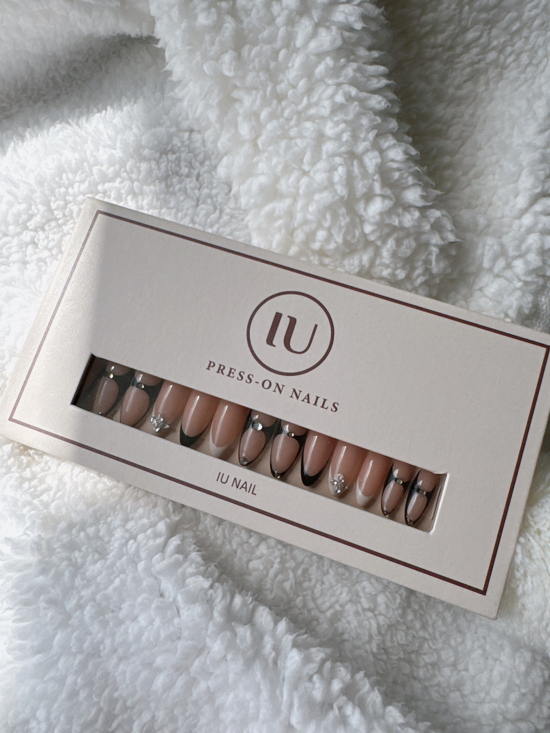 Nude press-on nails with black tips and rhinestone accents in a box on white plush fabric