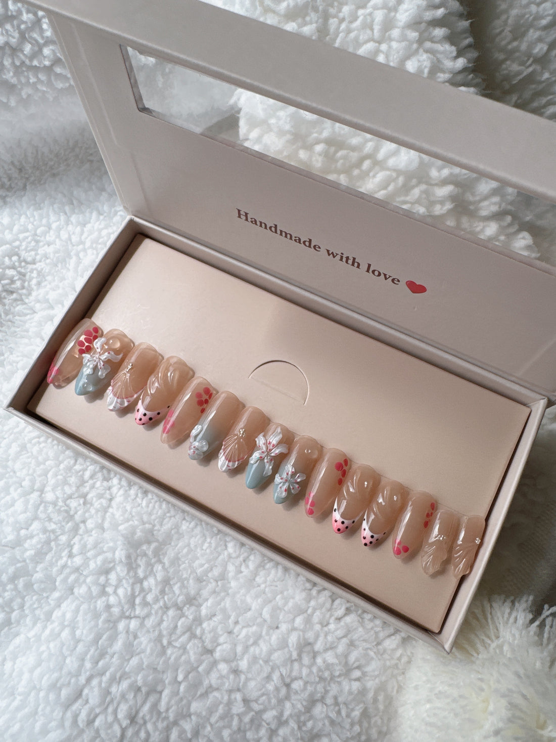 Box of colorful press-on nails with floral and polka dot designs on a white textured blanket.