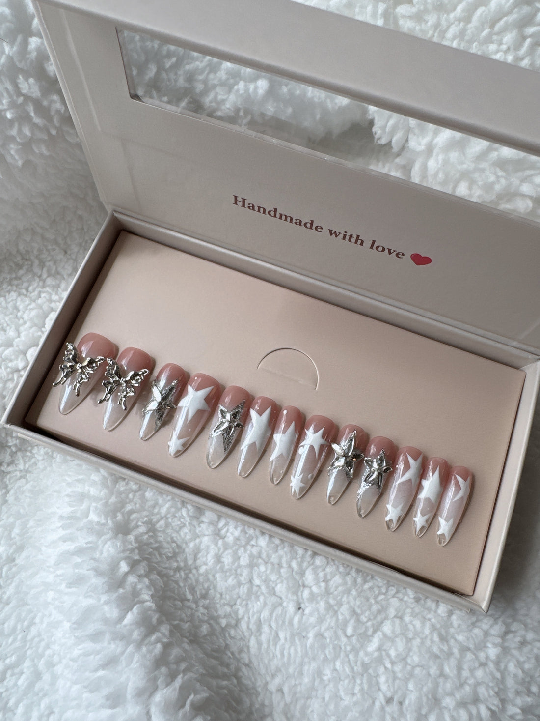 Press-on nails set with silver charms and star designs in pink packaging on a white textured blanket.