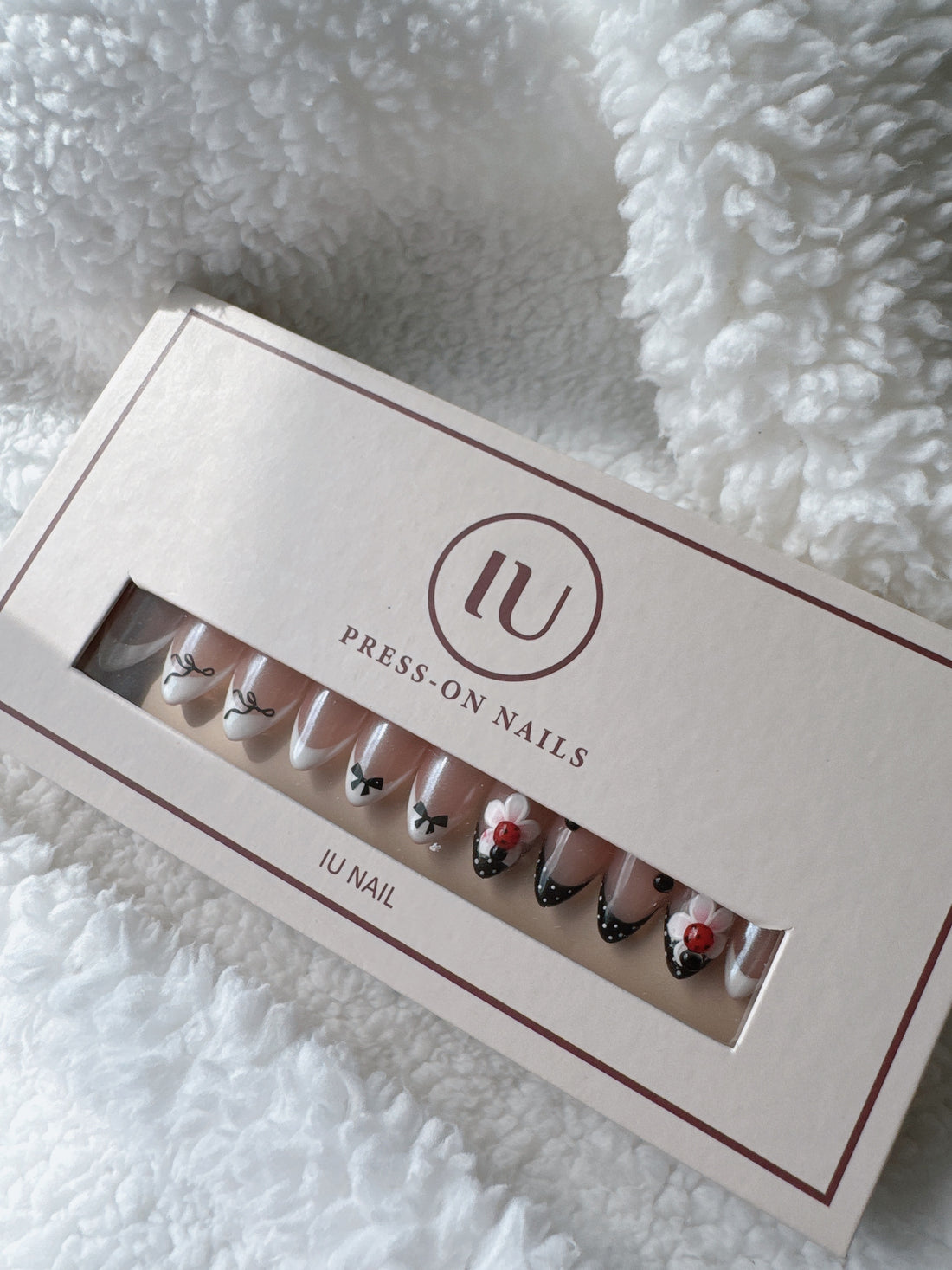 Press-on nails set with black bows, polka dots, and flower accents in box on white textured fabric.