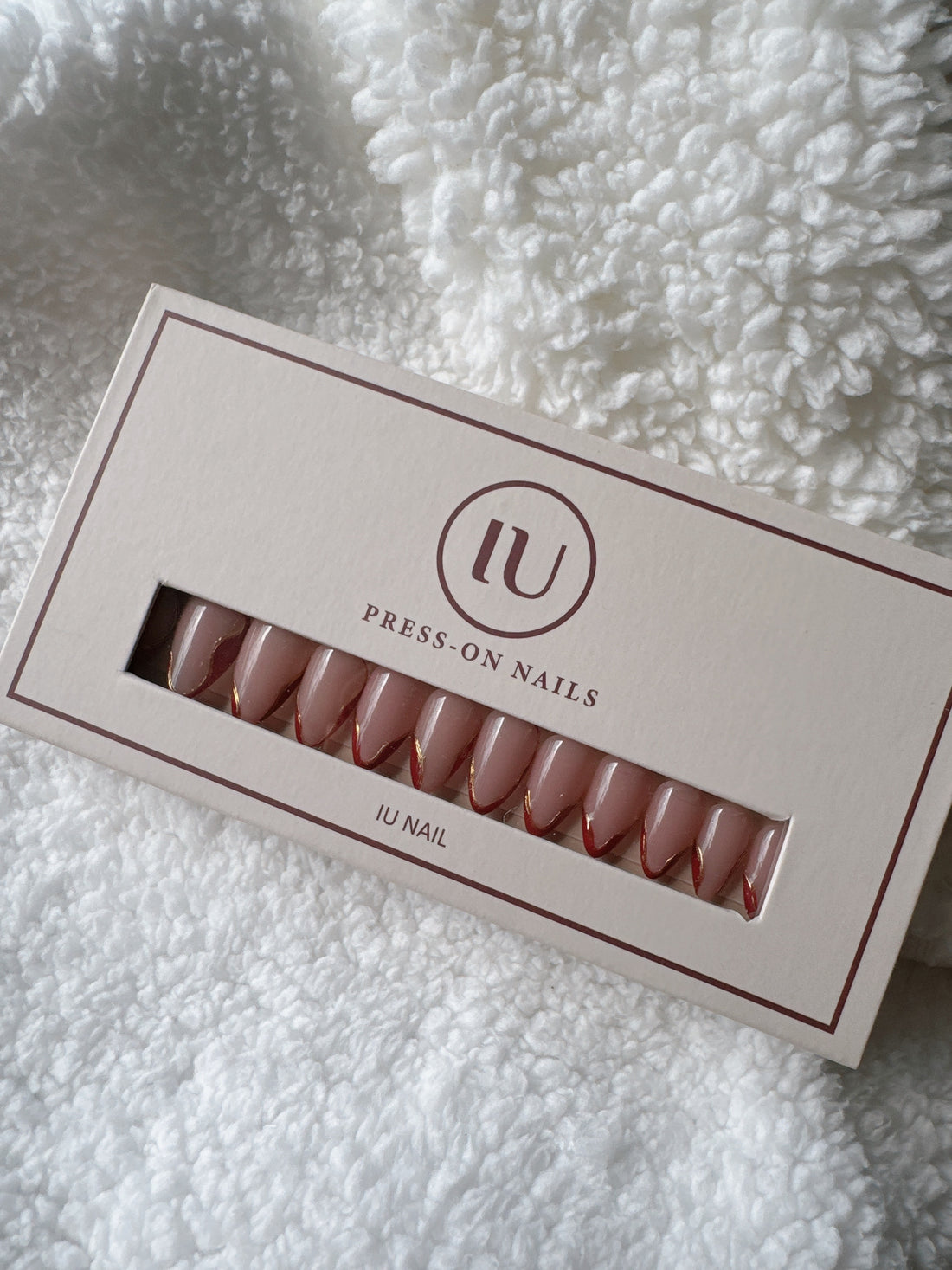 Box of nude almond-shaped press-on nails with gold and red tips on a white textured fabric
