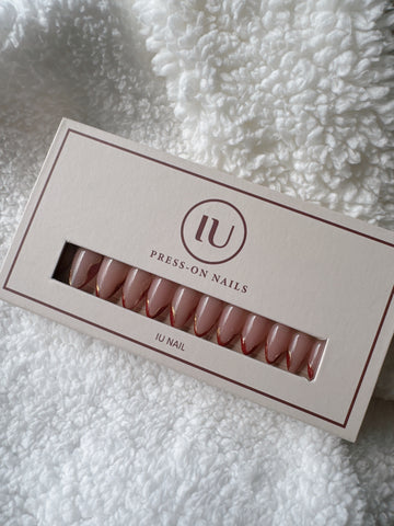 Box of nude almond-shaped press-on nails with gold and red tips on a white textured fabric