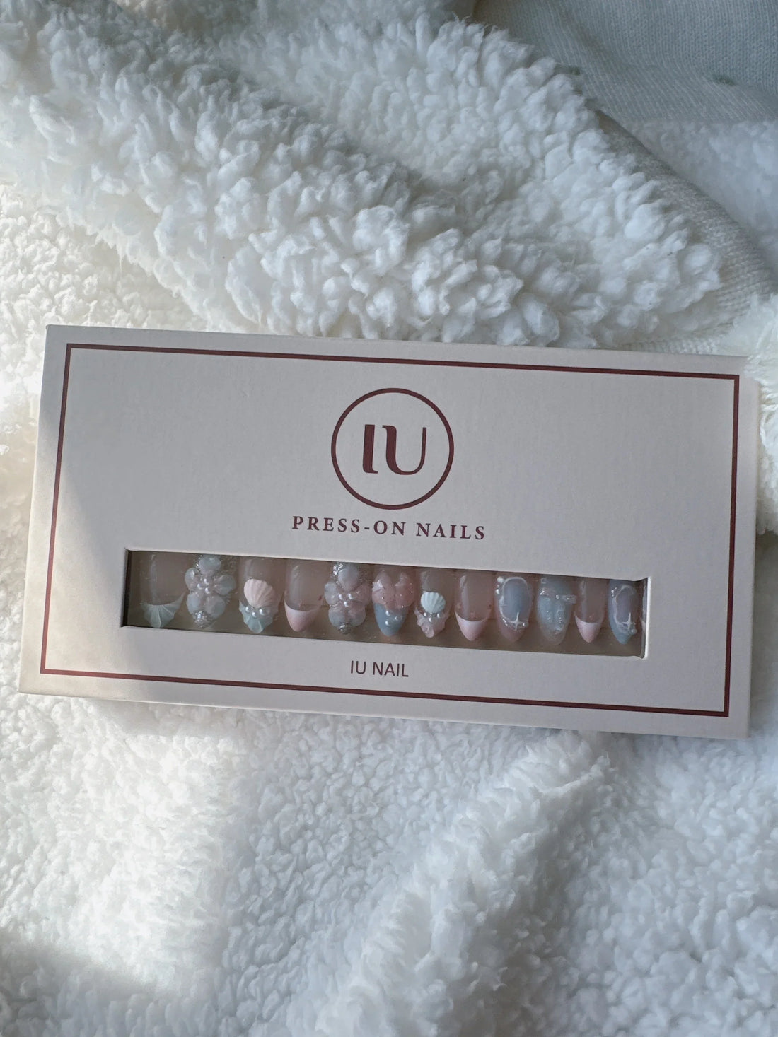 Box of pastel pink and blue press-on nails with 3D floral designs on soft white fabric.
