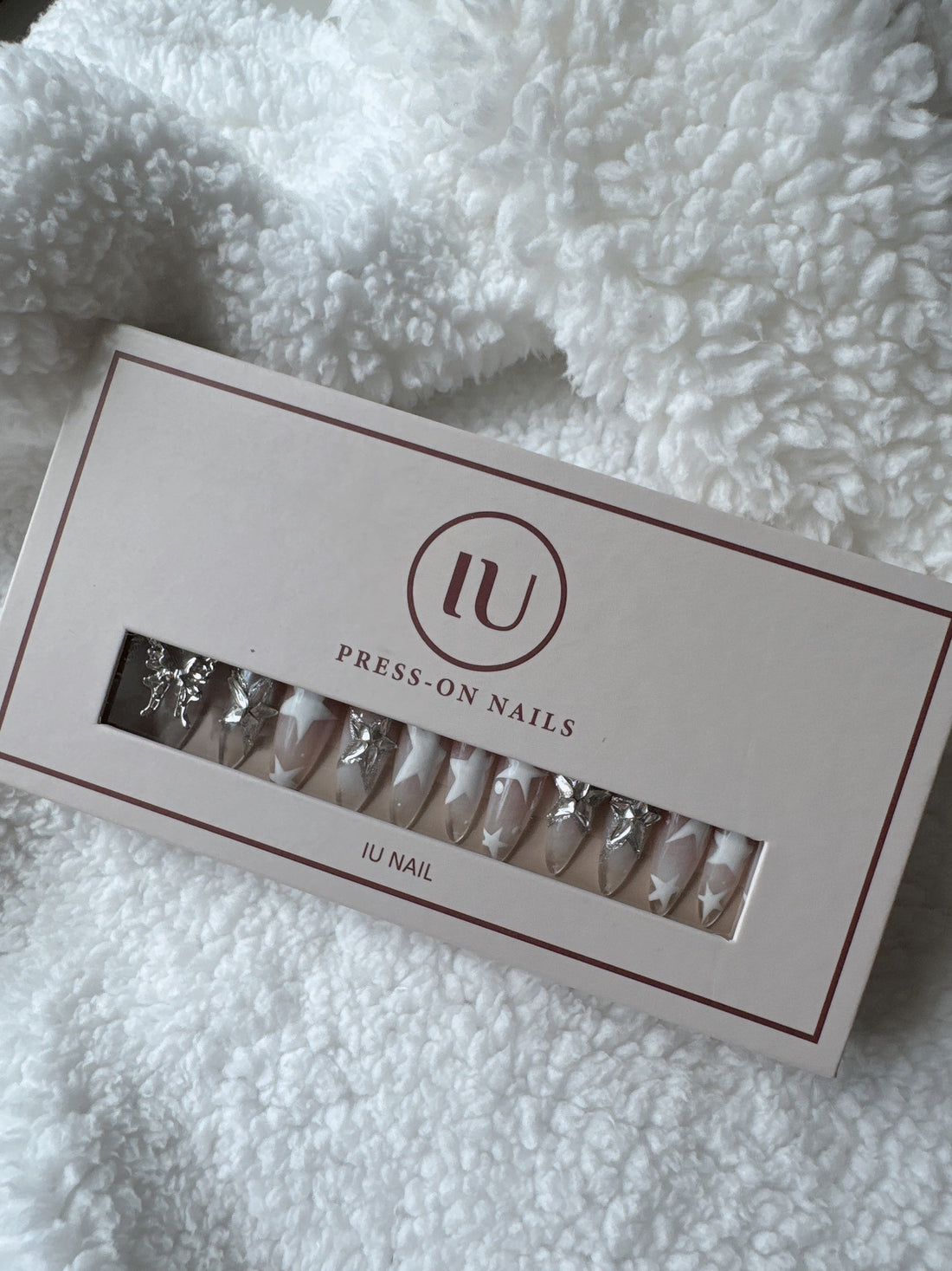Press-on nails set with silver charms and star designs in pink packaging on a white textured blanket.