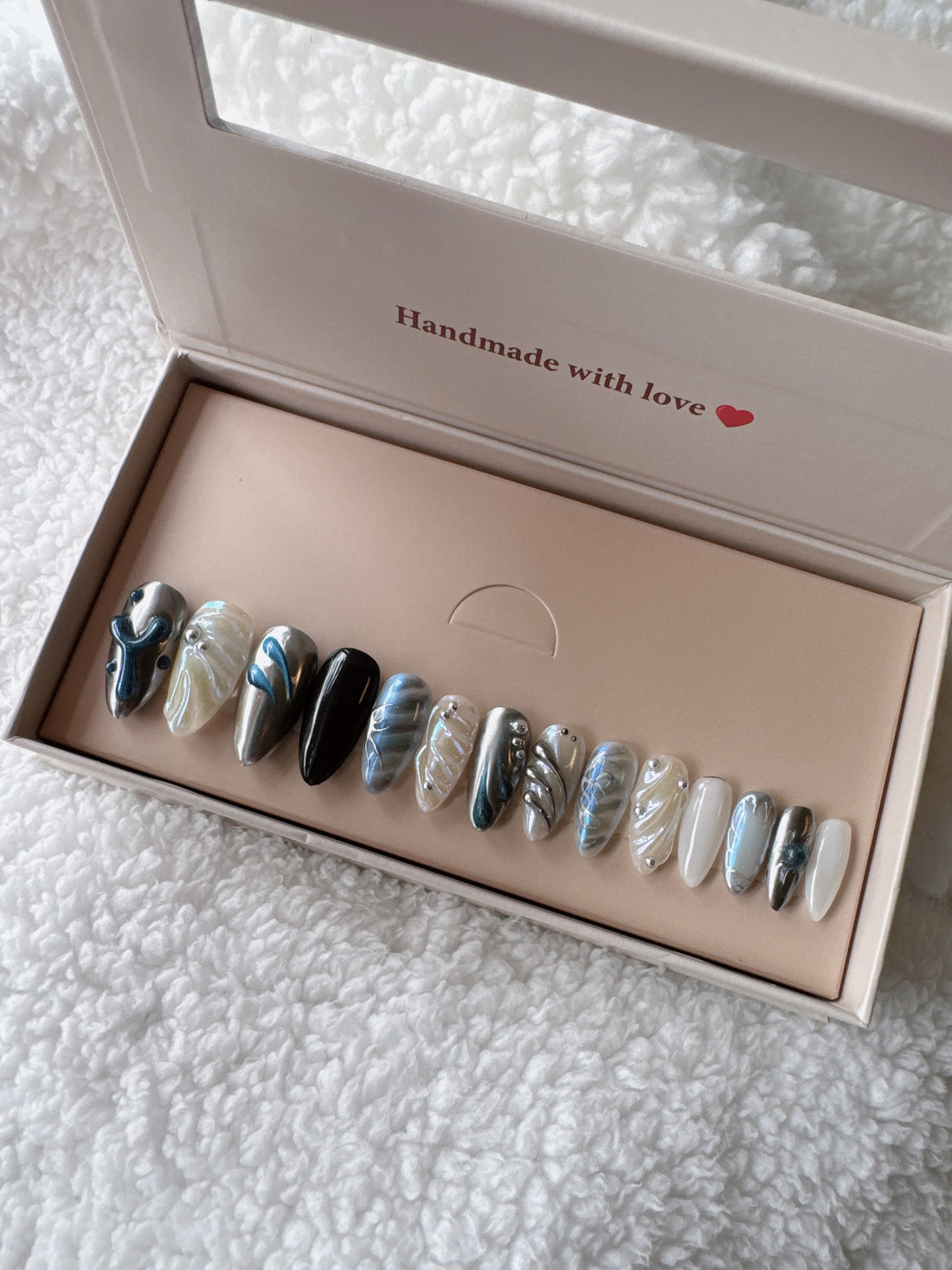 Box of IU press-on nails with marble and metallic designs on a white fluffy fabric