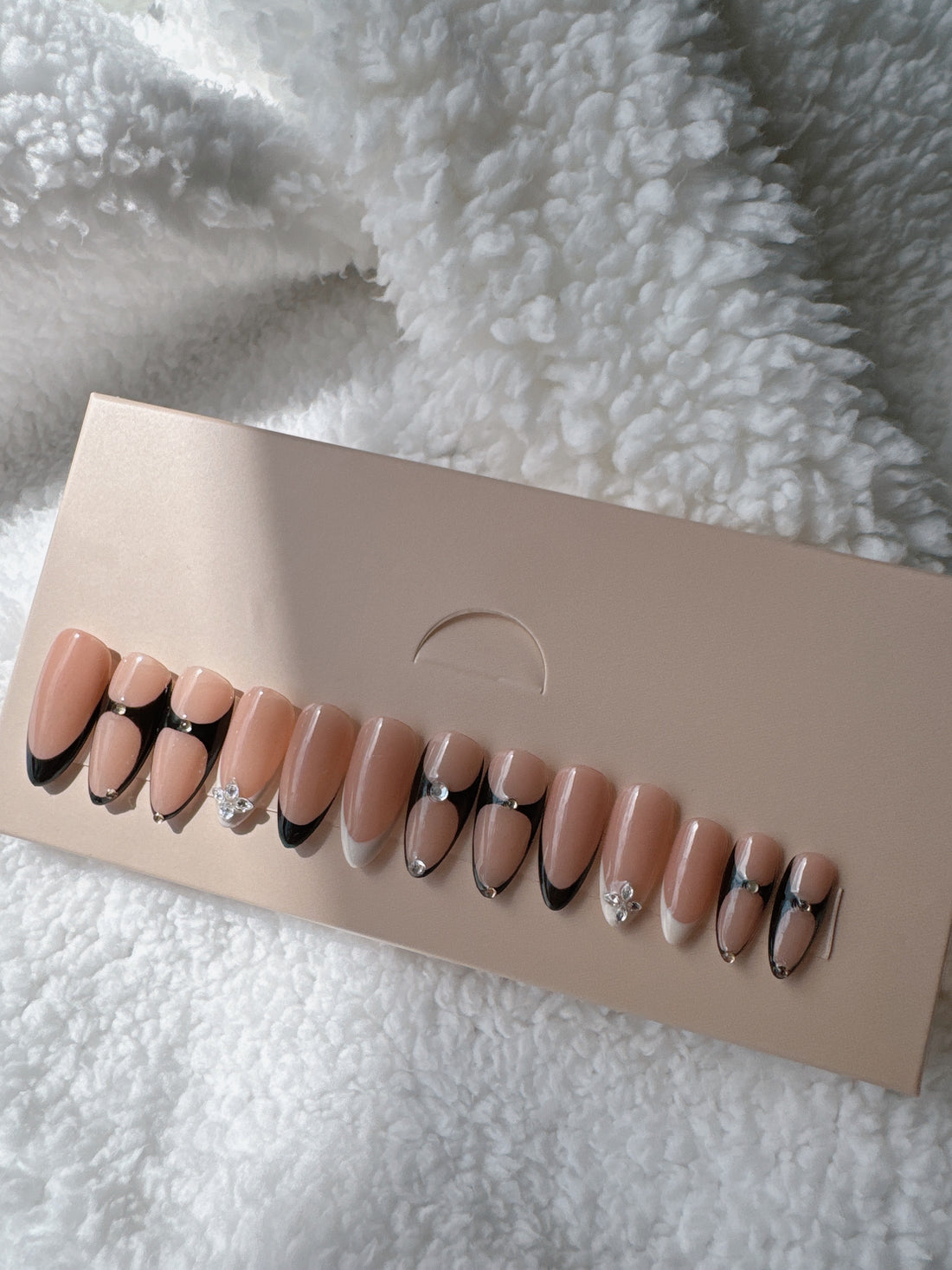 Nude press-on nails with black tips and rhinestone accents in a box on white plush fabric