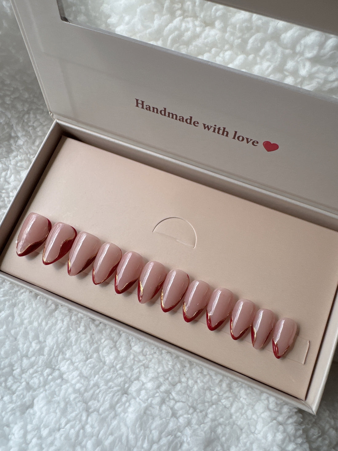 Box of nude almond-shaped press-on nails with gold and red tips on a white textured fabric