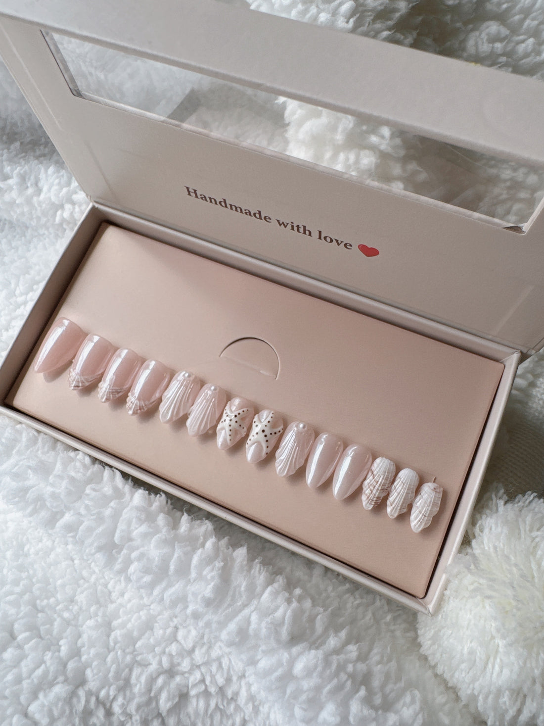 Box of nude and white press-on nails with delicate designs on soft white blanket